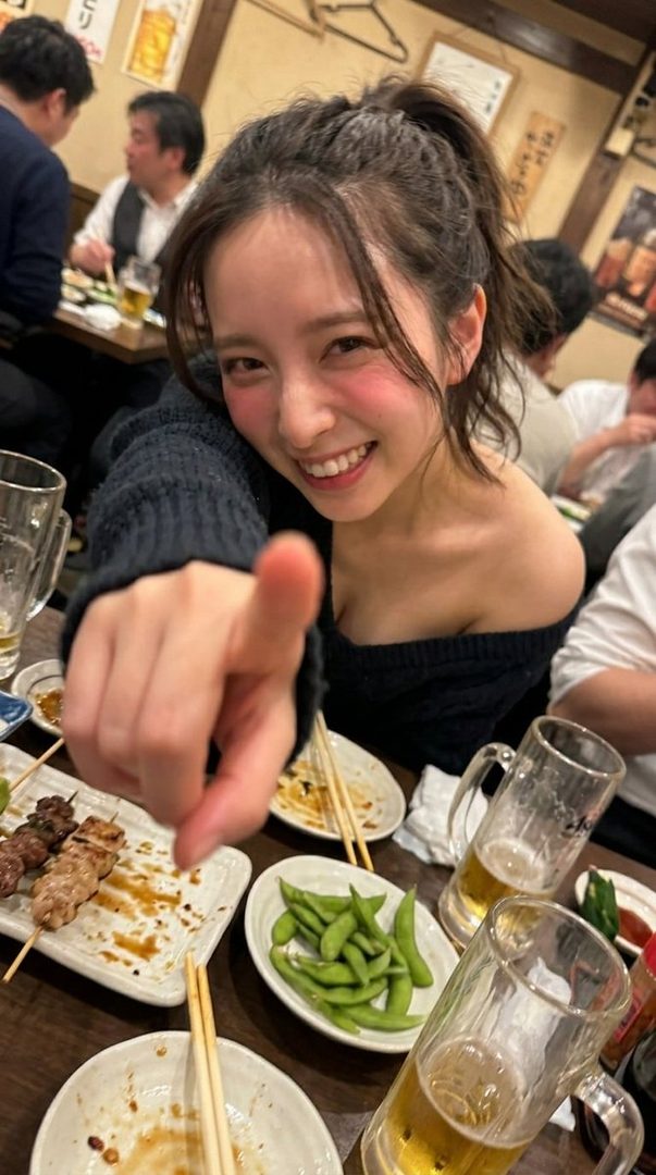 A young woman with her hair in a ponytail smiles and points at the camera while sitting at a table with food and drinks.
