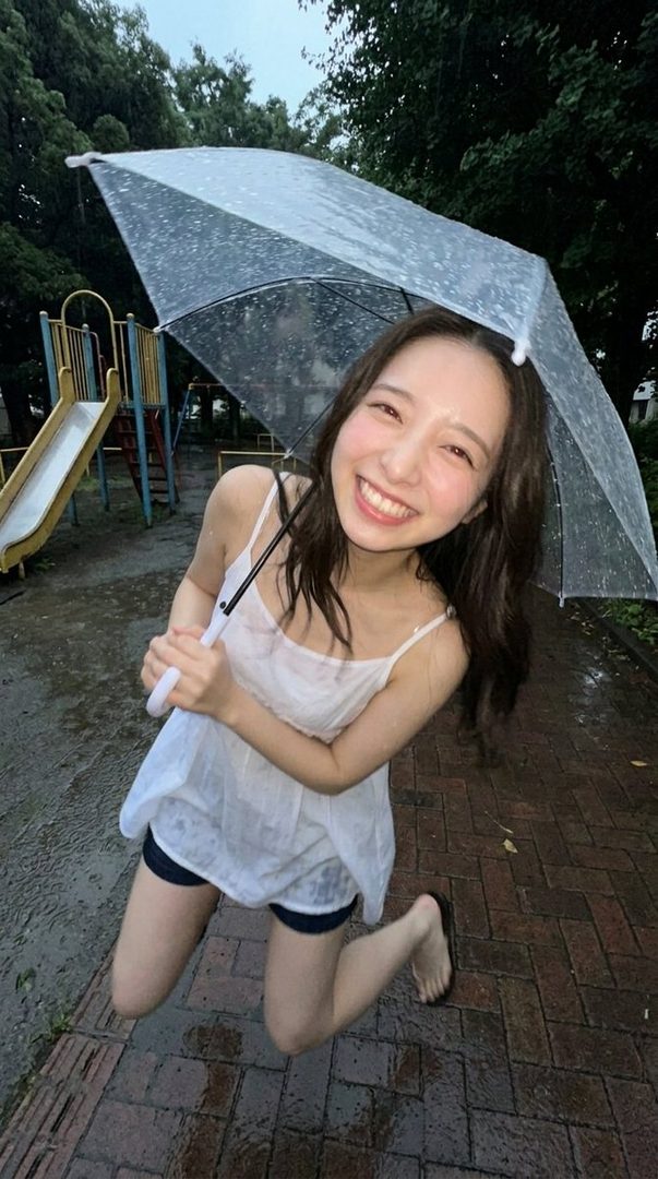 A young woman with long brown hair smiles brightly while holding a clear umbrella in the rain at a park playground.