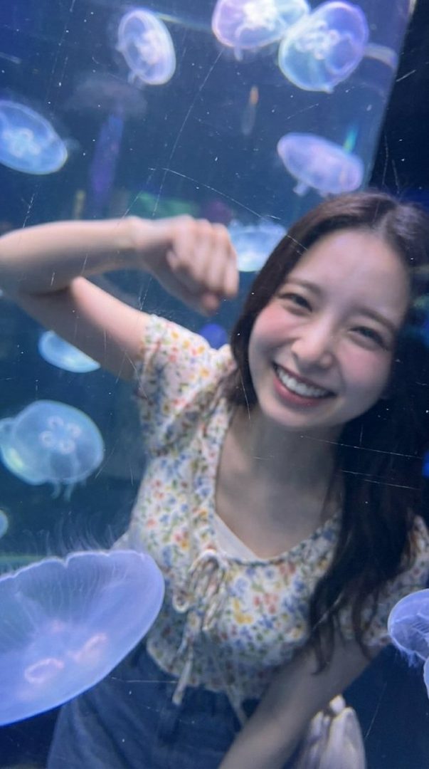 A young woman with long brown hair smiles brightly while posing in front of a jellyfish tank at an aquarium.