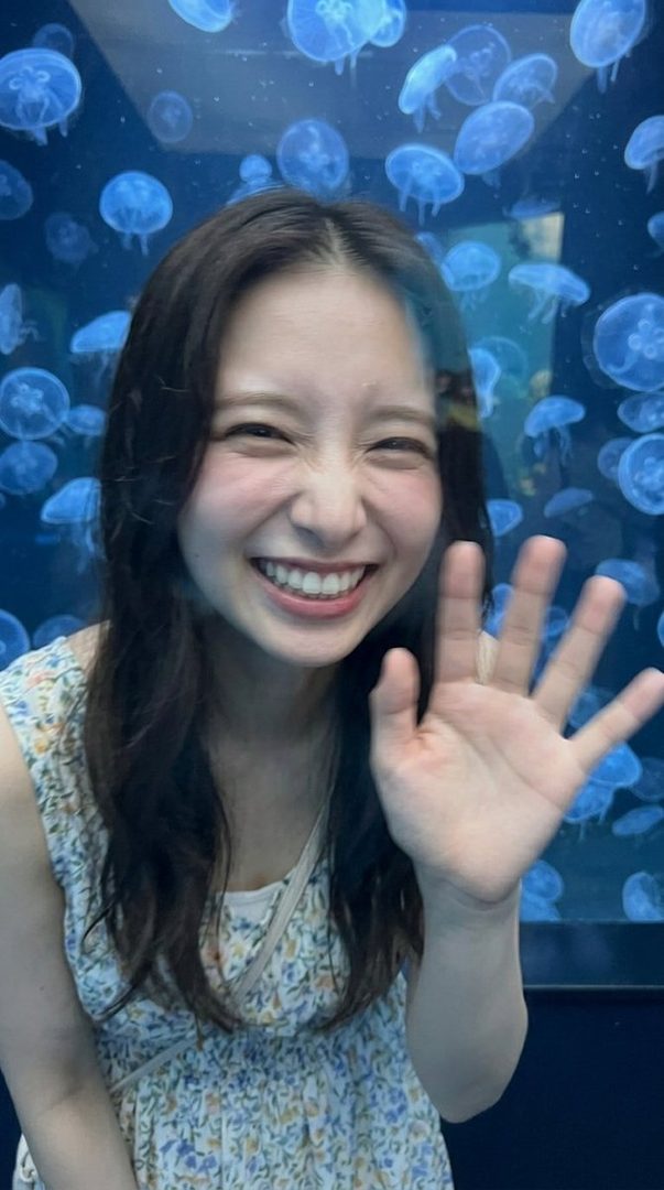 A young woman with long brown hair smiles and waves at the camera in front of a jellyfish exhibit at an aquarium.
