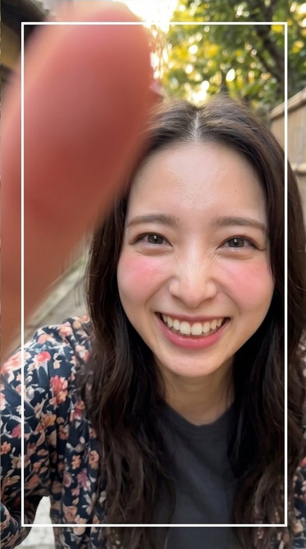 A young woman with long brown hair smiles brightly at the camera, her cheeks flushed with pink blush, wearing a floral print top.