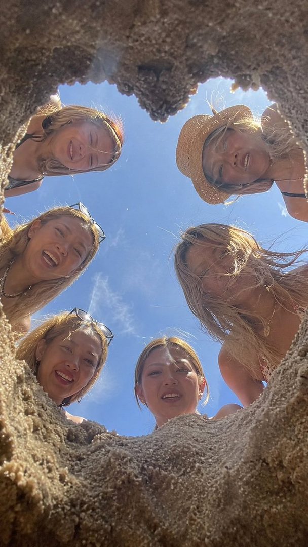 A group of five young women with blonde hair smile down at the camera from a beach.