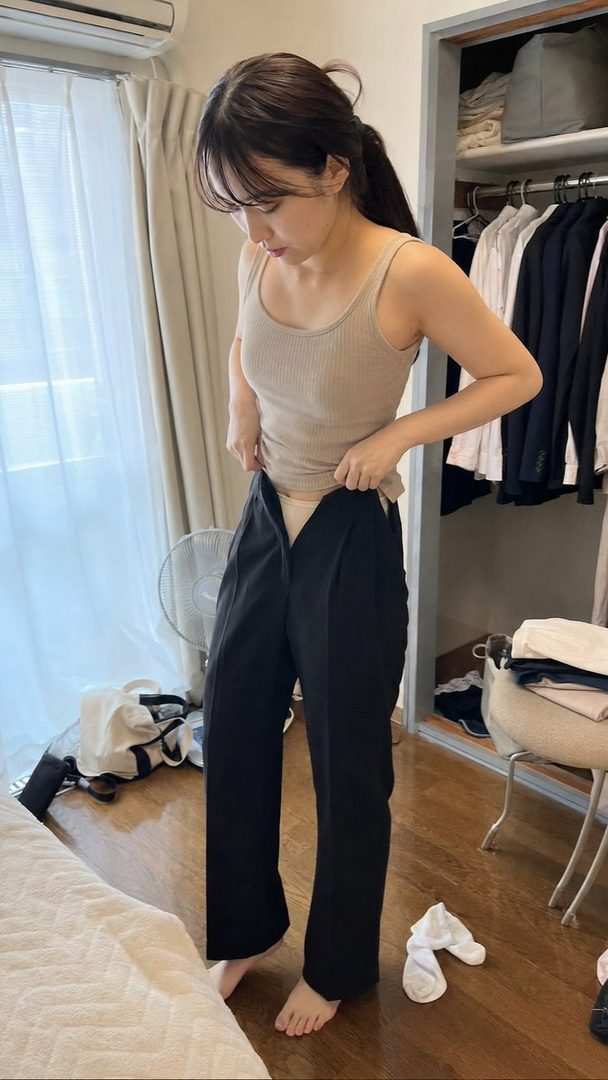 A young woman with her hair in a ponytail is getting dressed in her bedroom, with a closet full of clothes in the background.