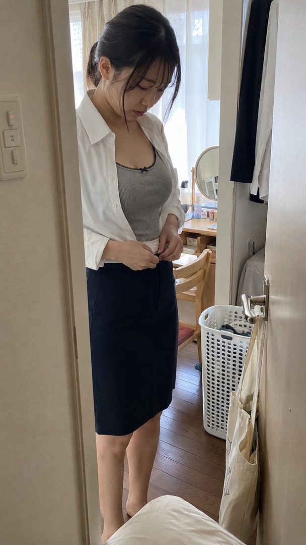 A young woman in a white shirt and grey tank top, adjusting her skirt in a room.