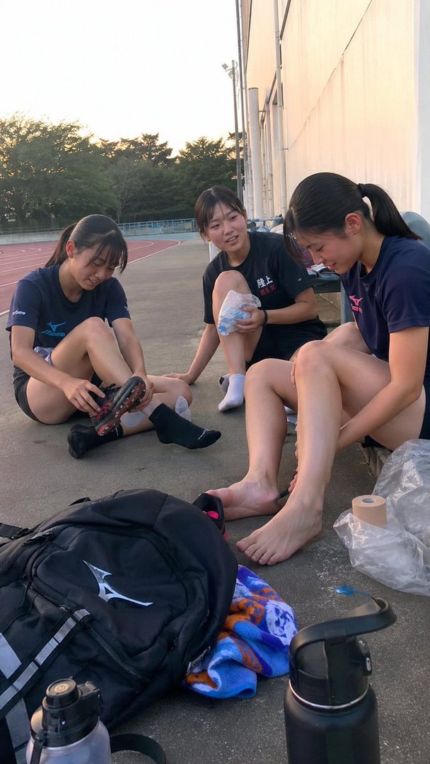 Three young female athletes resting after a sports practice, sitting on the ground near a track and field stadium.