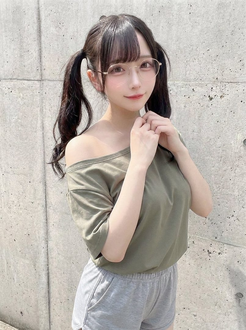 Young woman with black twin pigtails and thin glasses wearing an off-shoulder olive top and gray sweatpants posing against a concrete wall.