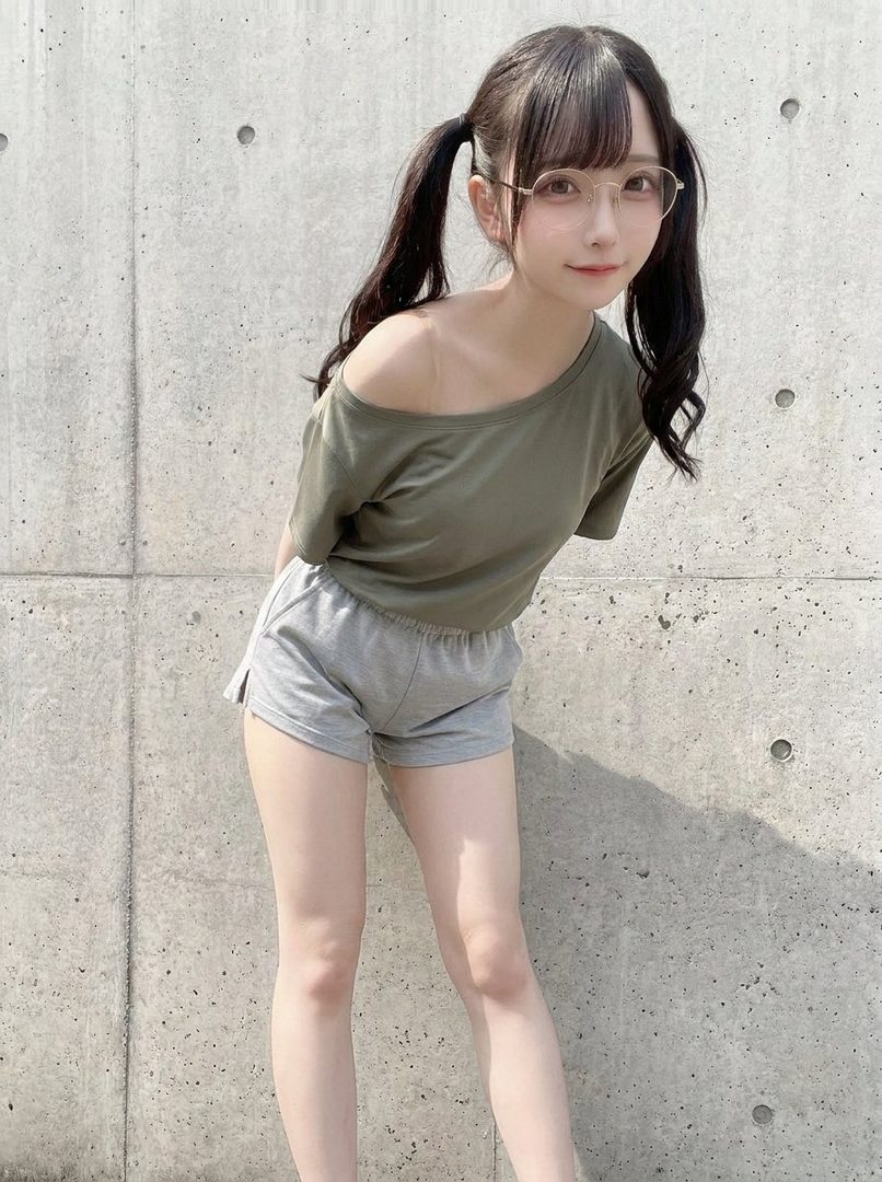 A young woman with black twintails and glasses posing casually in an olive top and gray shorts against a concrete wall.