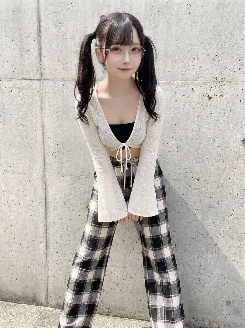 Young Japanese woman with black pigtails and glasses posing in sparkly white crop top and plaid pants against a concrete wall.