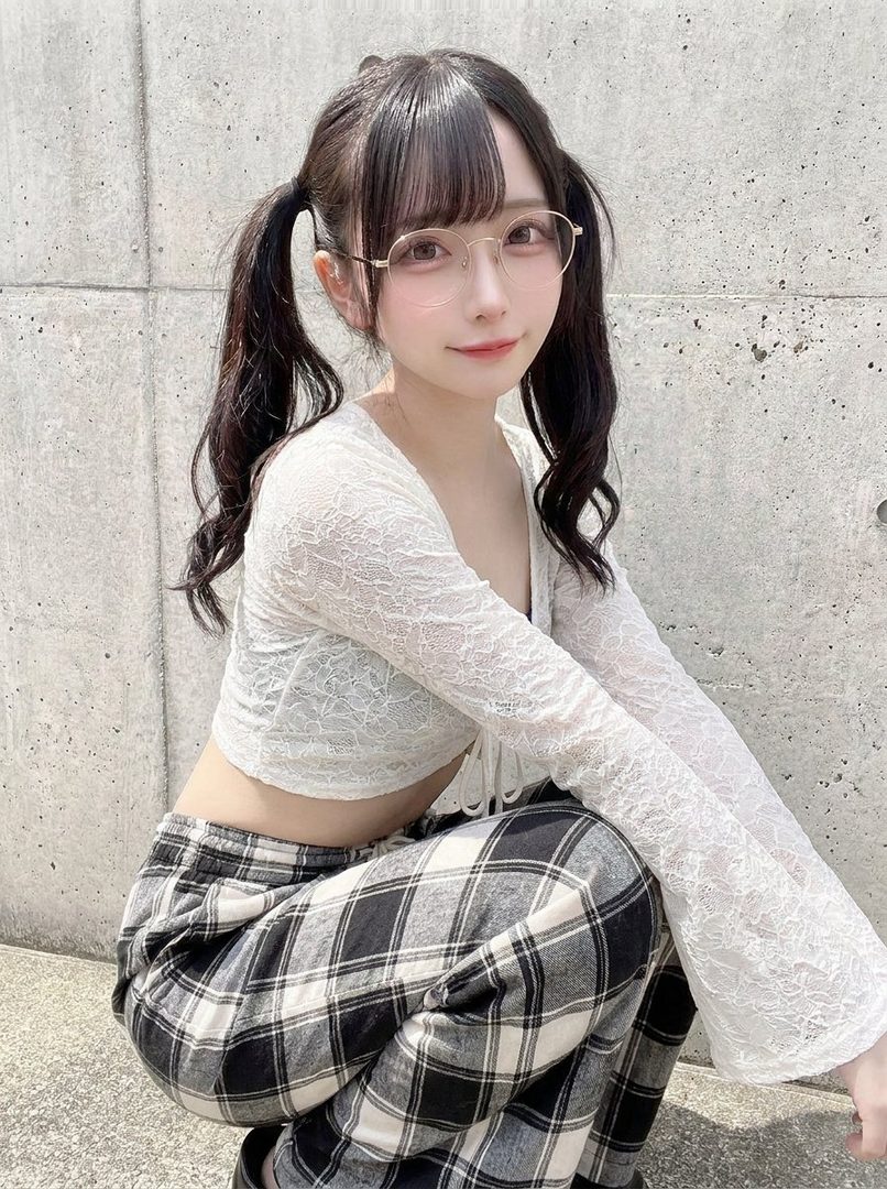 A young woman with black hair in pigtails and glasses, wearing a white lace top and plaid pants, posing outdoors.