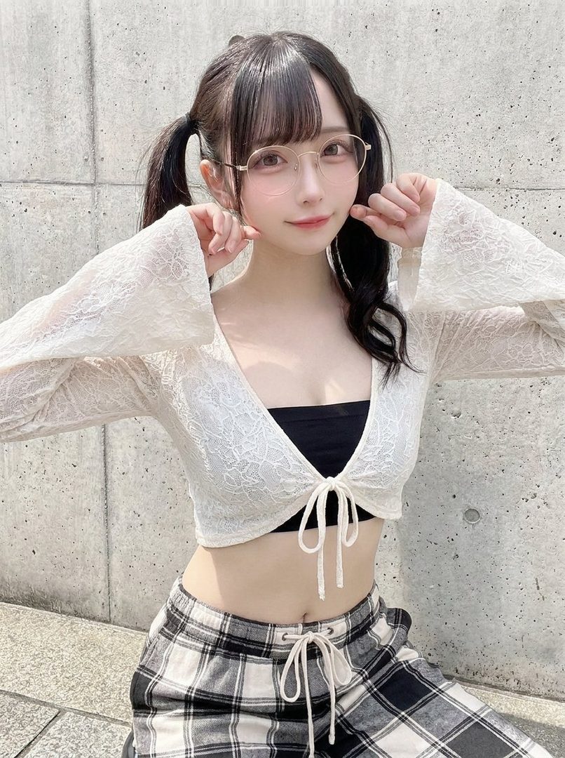 A young woman with black hair in pigtails and bangs, wearing glasses, a white lace top, and plaid pants, poses outdoors.