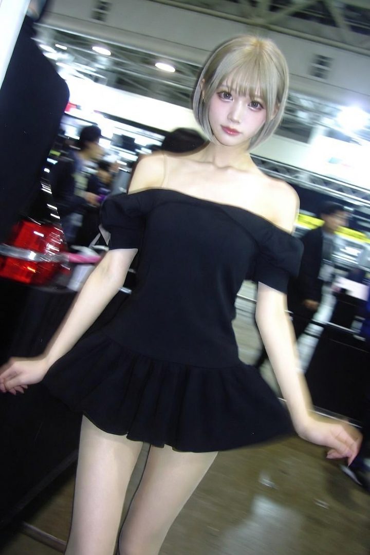 A young woman with short blonde hair and bangs, wearing an off-the-shoulder black dress, poses indoors.