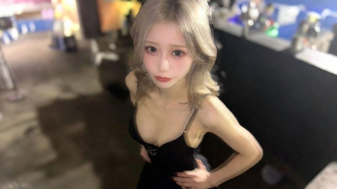 A young woman with blonde hair wearing a black dress, posing indoors.