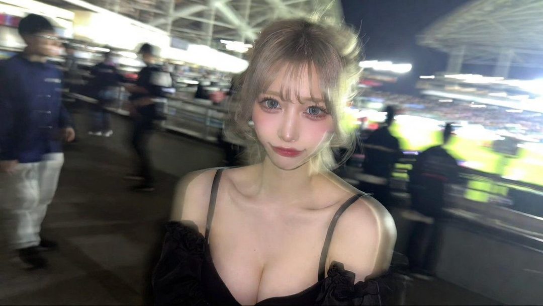A young woman with blonde hair and striking makeup poses for a photo at a stadium at night.