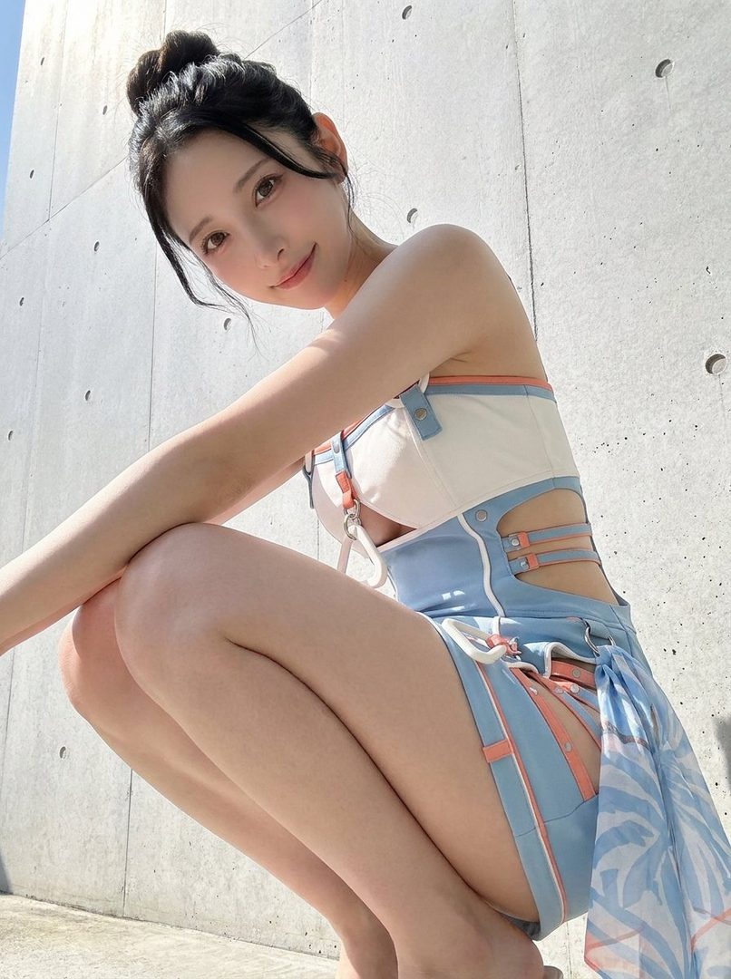 A young woman with her hair in a bun, wearing a blue and white outfit, poses outdoors against a concrete wall.