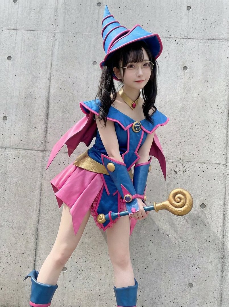 A young woman in a blue and pink Dark Magician Girl costume from Yu-Gi-Oh!, complete with a pointed hat and staff, posing outdoors.
