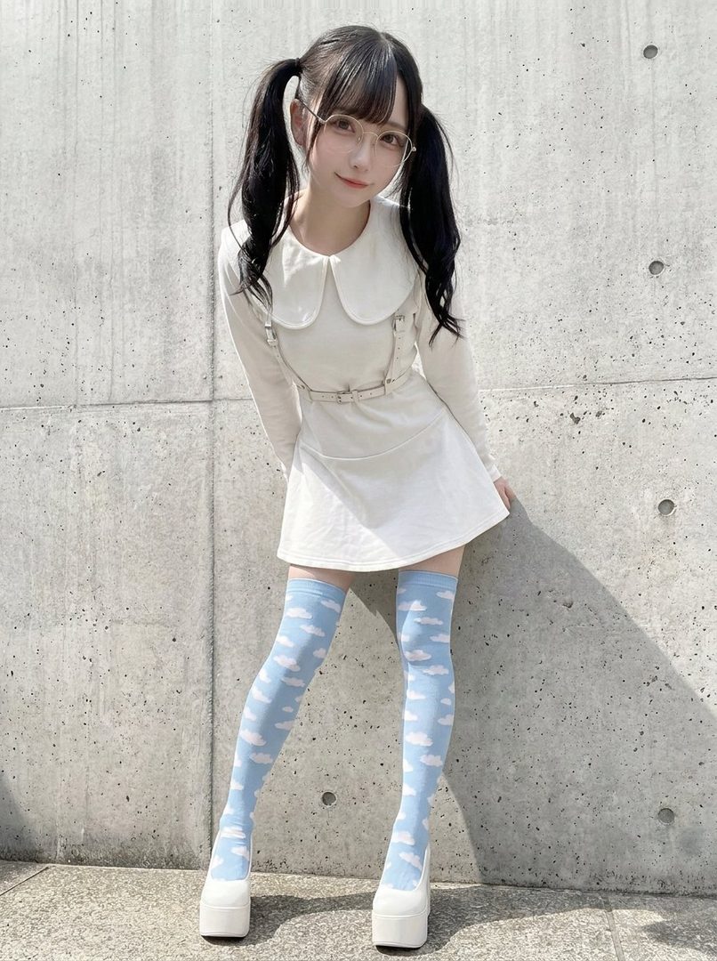 A young woman with black twin-tail hair and glasses, wearing a white dress and thigh-high socks with a cloud pattern, poses outdoors.