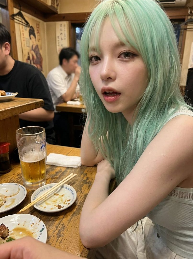 A young woman with long, mint-green hair and bangs, wearing a light-colored top, poses for a portrait in an indoor setting with a glass of beer and food on the table.