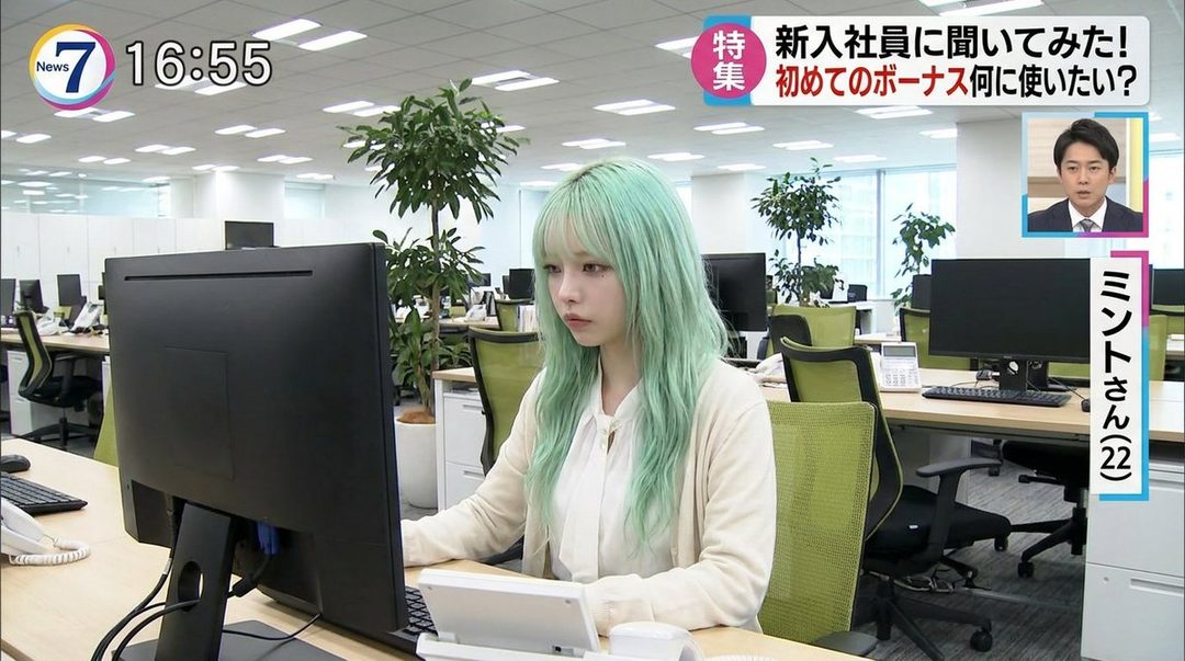 A young woman with long, mint green hair and bangs, wearing a white blouse and a cream cardigan, sits at a desk in a modern office, looking at a computer screen.