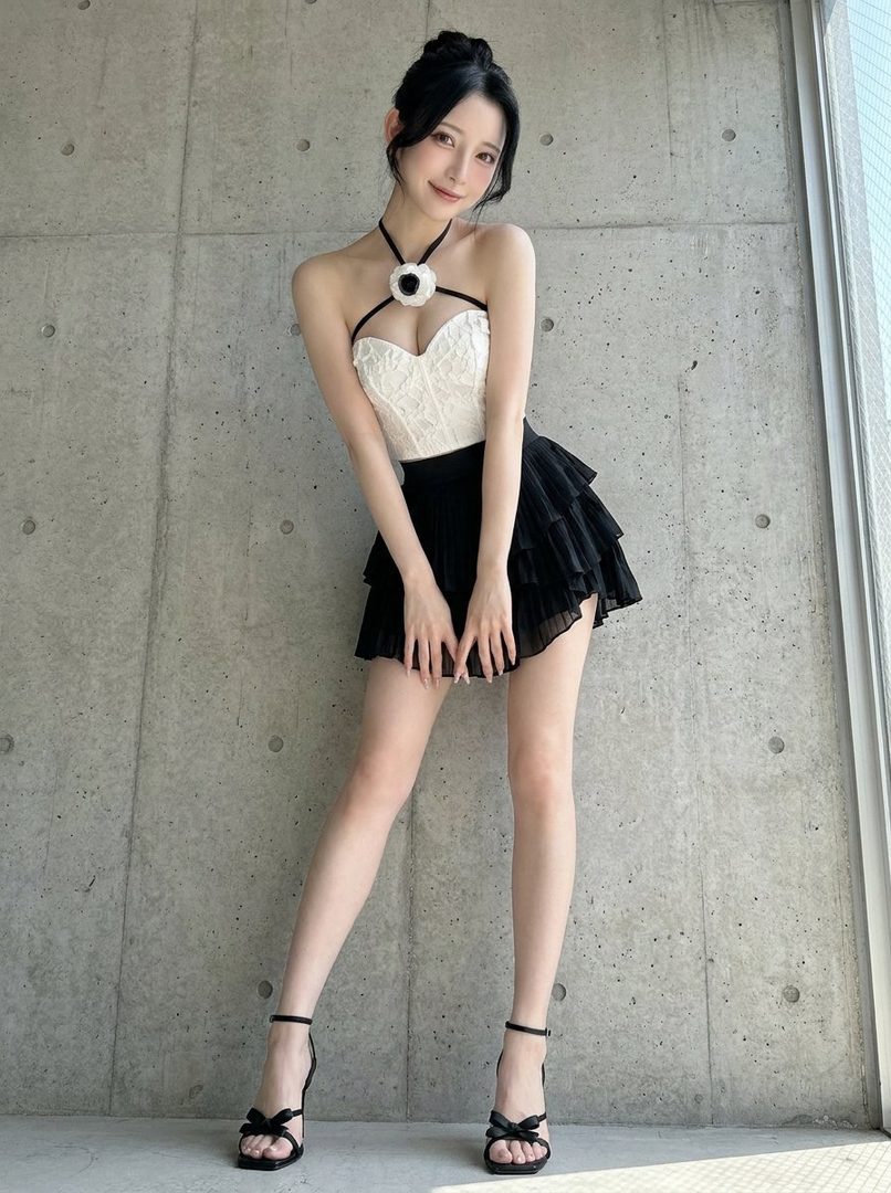 A young woman with black hair in a bun, wearing a white lace corset top and a black ruffled skirt, posing indoors.