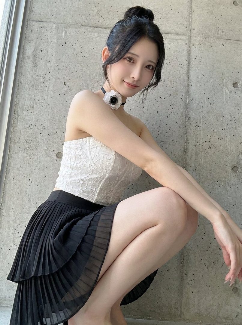 A young woman with her hair in a bun, wearing a white lace strapless top and a black pleated skirt, poses indoors.
