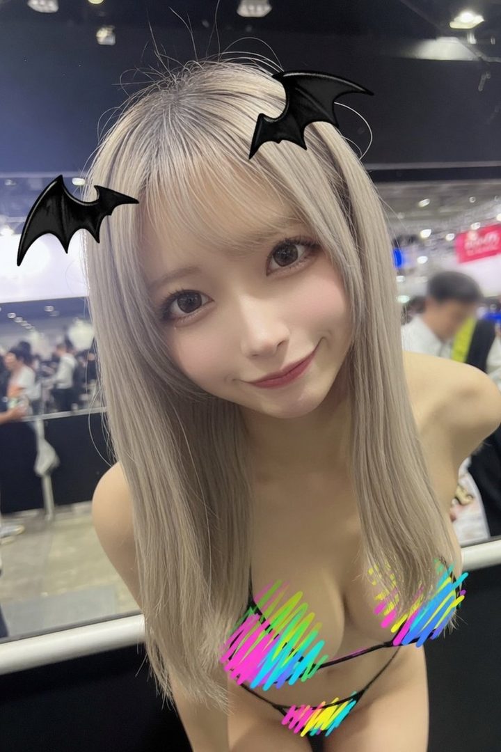 A young woman with long blonde hair and bangs, wearing a colorful bikini top, smiles at the camera with bat decorations in her hair.