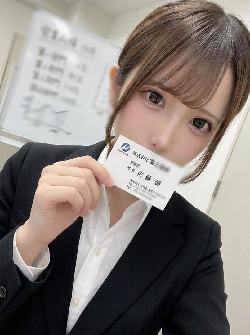 A woman in a black suit and white shirt holds up a business card, obscuring her mouth. She has brown hair with bangs and is looking at the camera.