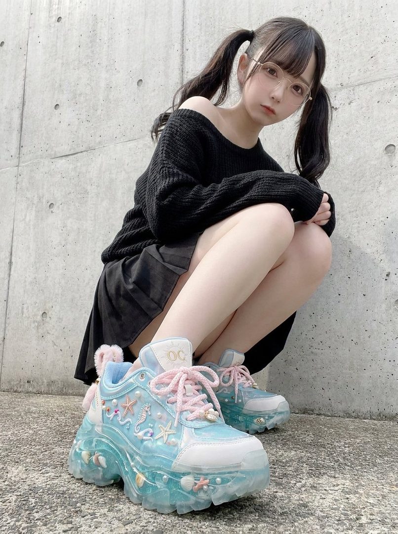 A young woman with black pigtails and glasses, wearing a black off-the-shoulder sweater and a short black skirt, crouches outdoors in front of a concrete wall, showcasing her unique platform sneakers decorated with seashells and seahorses.