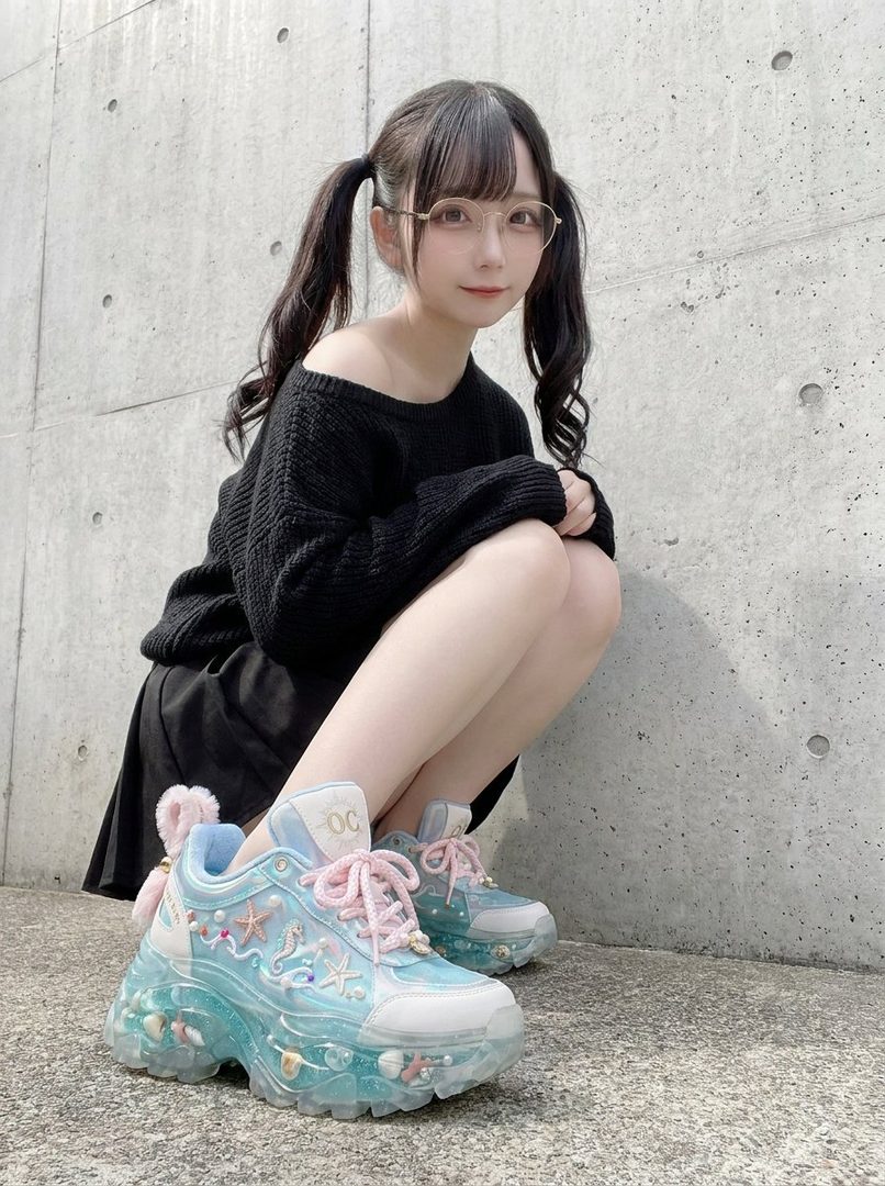 A young woman with black pigtails and glasses, wearing a black off-the-shoulder sweater and a black skirt, crouches down in front of a concrete wall, showcasing her unique blue sneakers decorated with seashells and starfish.