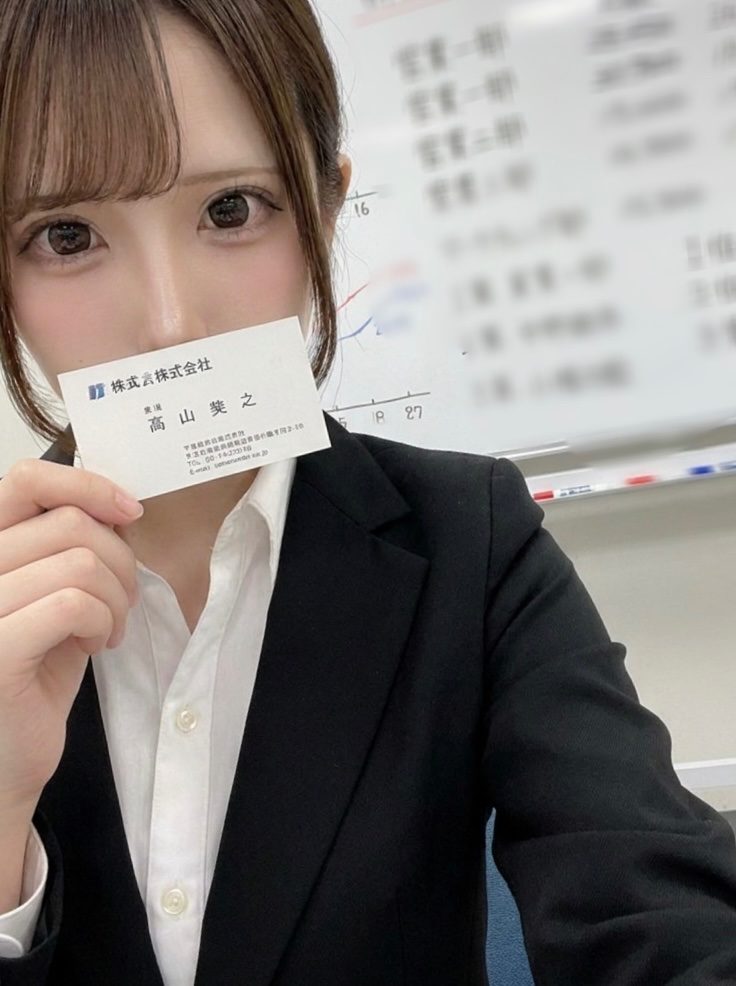 A young woman in a black suit and white shirt holds up a business card in front of her face, with a whiteboard in the background.