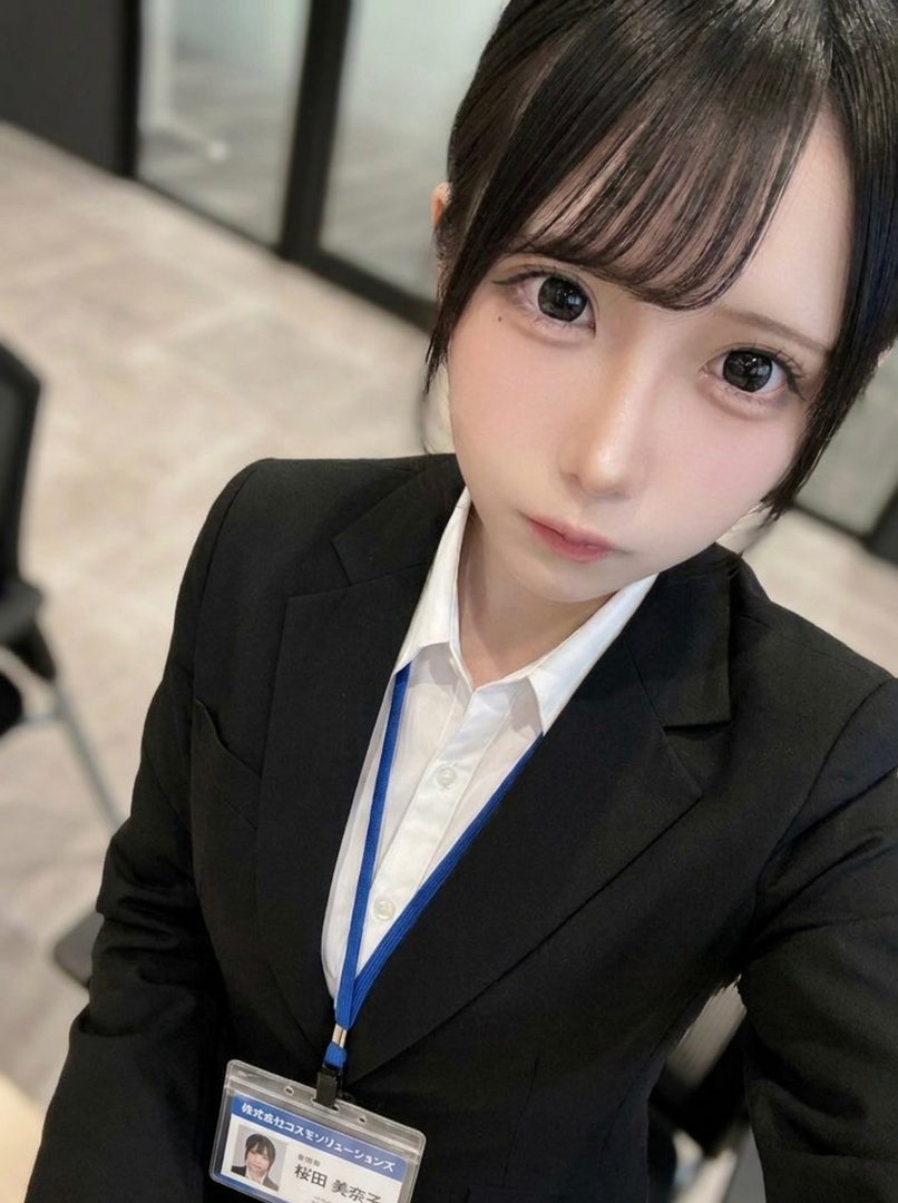 A young woman in a black suit and white shirt, wearing an ID badge, poses for a selfie in an office setting.