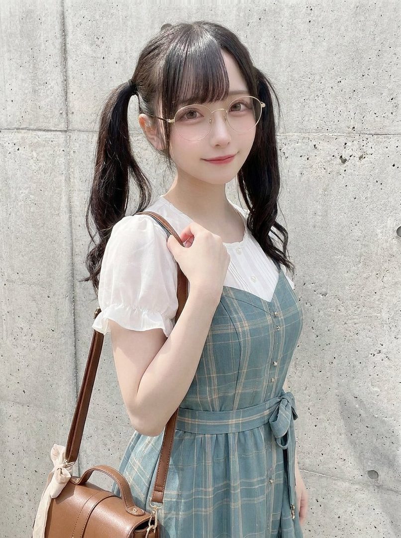 A young woman with black hair in pigtails and bangs, wearing glasses and a plaid dress, poses outdoors.