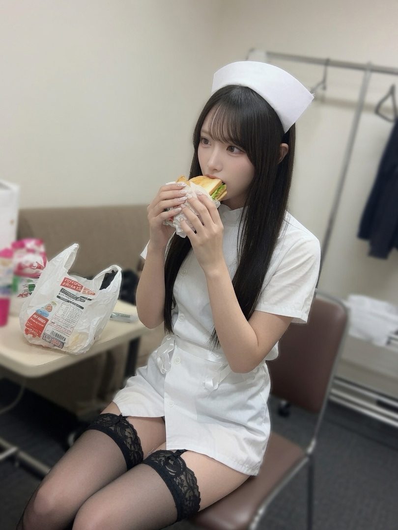 Young Japanese woman in white nurse cosplay uniform eating a sandwich indoors.