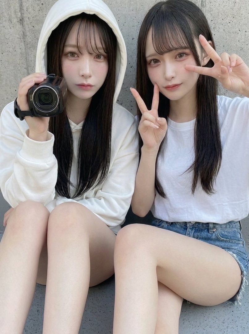 Two young women with long black hair and similar features posing together against a concrete wall, one holding a camera while the other makes peace signs.