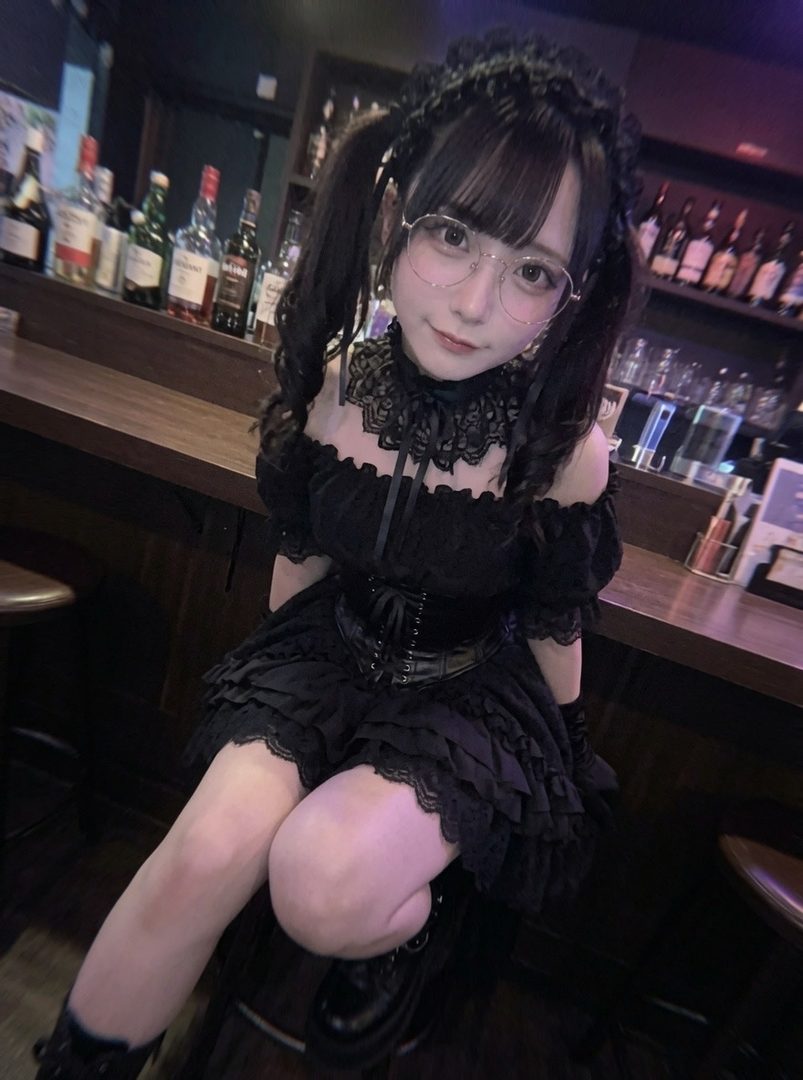Young woman in black gothic lolita dress with pigtails and glasses posing at a dimly lit bar counter.