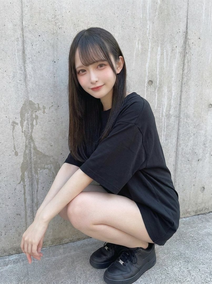 Young Japanese woman with long black hair and bangs squatting casually against a concrete wall in black oversized t-shirt and sneakers.
