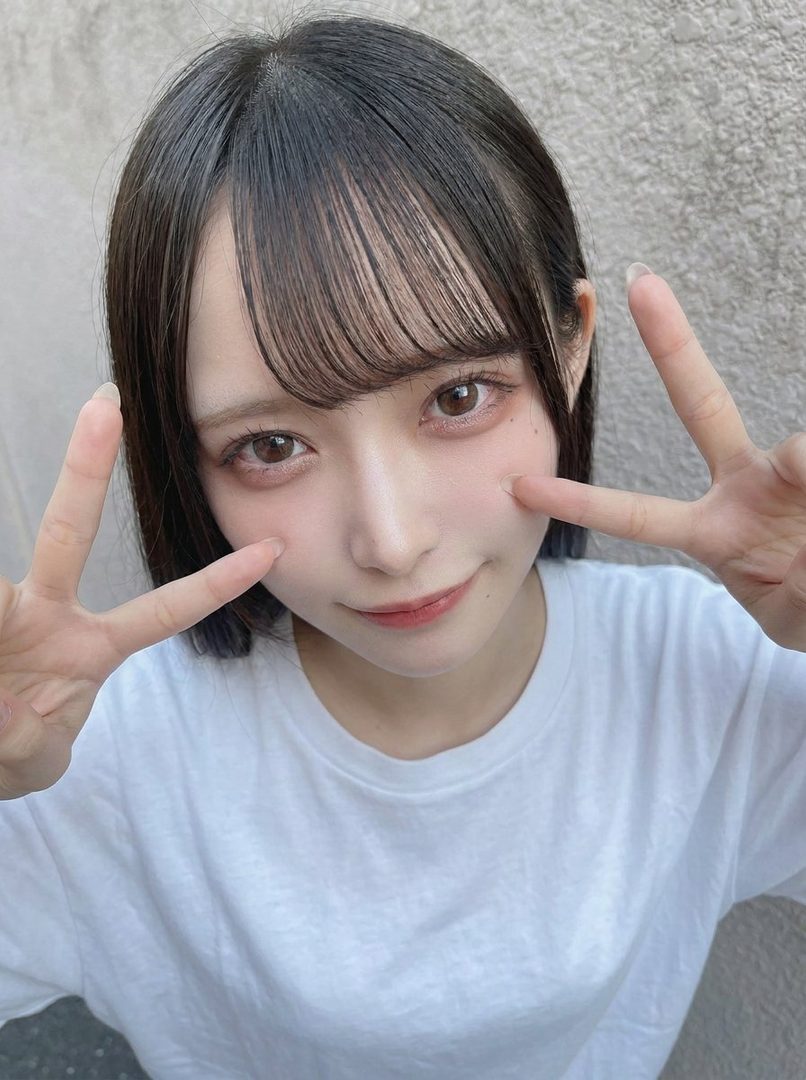 Young East Asian woman with black bob haircut and bangs taking a close-up selfie while making playful peace signs against a textured wall.