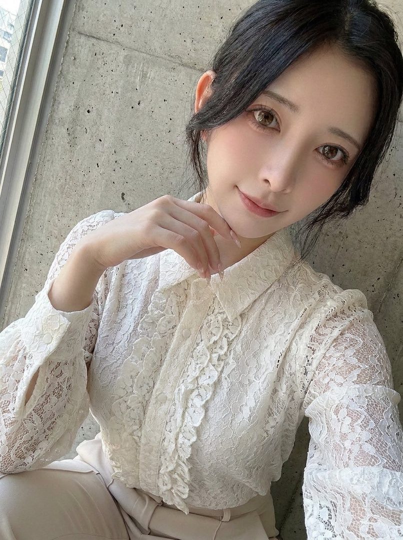 A young East Asian woman with long dark hair wearing a white lace blouse poses elegantly indoors against a concrete wall.
