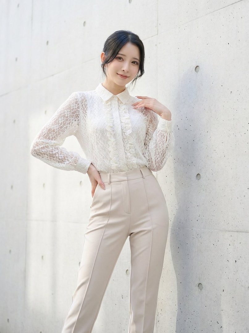 Elegant woman with dark hair wearing a white lace blouse and beige trousers posing against a concrete wall.