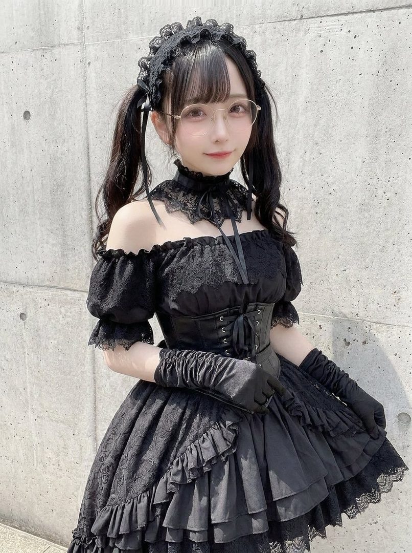 Young woman in an elaborate black gothic lolita dress with twin tails, glasses, and lace accessories posing outdoors against a concrete wall.
