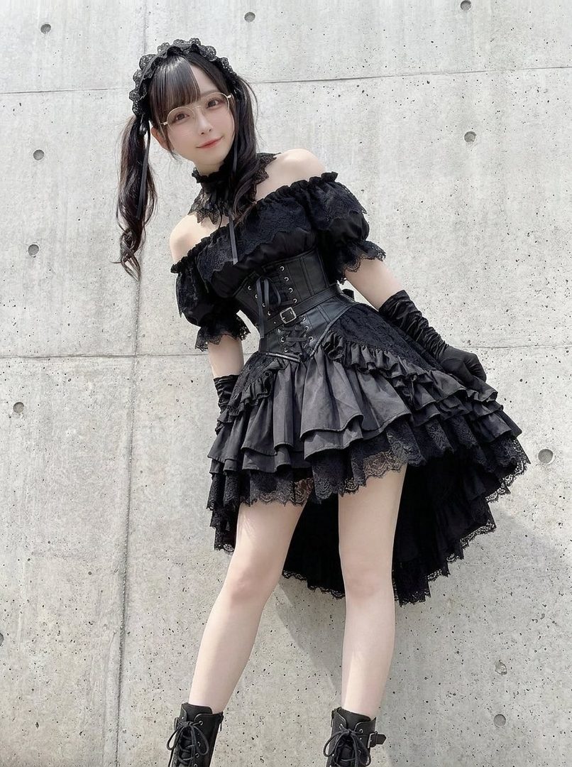 Young woman in elaborate black gothic lolita dress with corset and ruffles posing against a concrete wall.