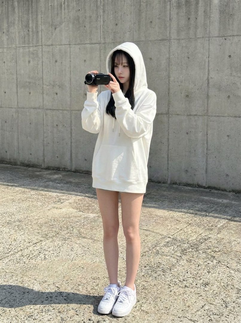 Young woman in oversized white hoodie holding a camcorder while standing against a concrete wall outdoors.