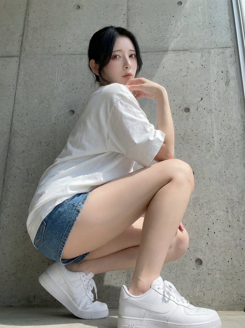 Young East Asian woman with long black hair posing in an oversized white t-shirt and denim shorts against a concrete wall.