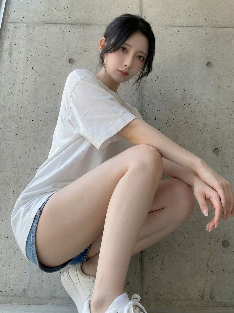Young East Asian woman with long black hair posing seated against a concrete wall in a loose white t-shirt and denim shorts, highlighting her long legs.