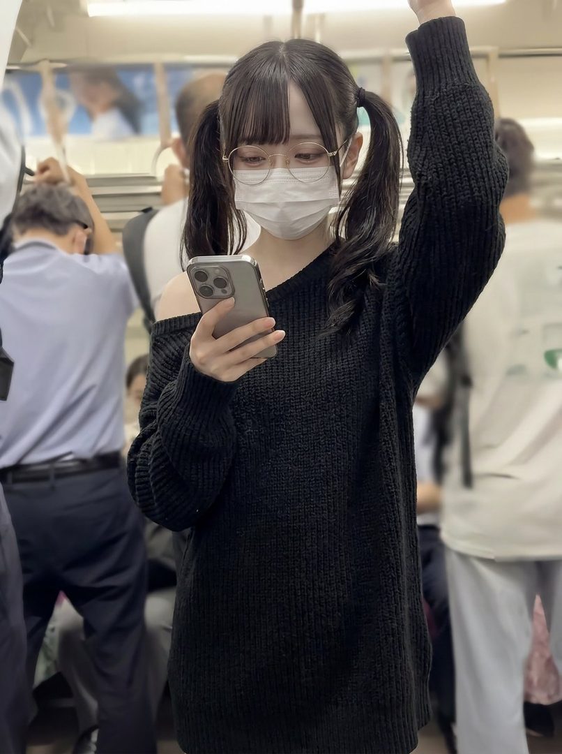 Young woman with black twintails taking a selfie on a train while wearing glasses, a face mask, and an oversized black knit sweater.