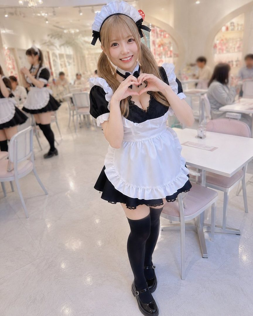 Smiling blonde woman in a black and white maid outfit posing with a heart gesture inside a bright cafe.