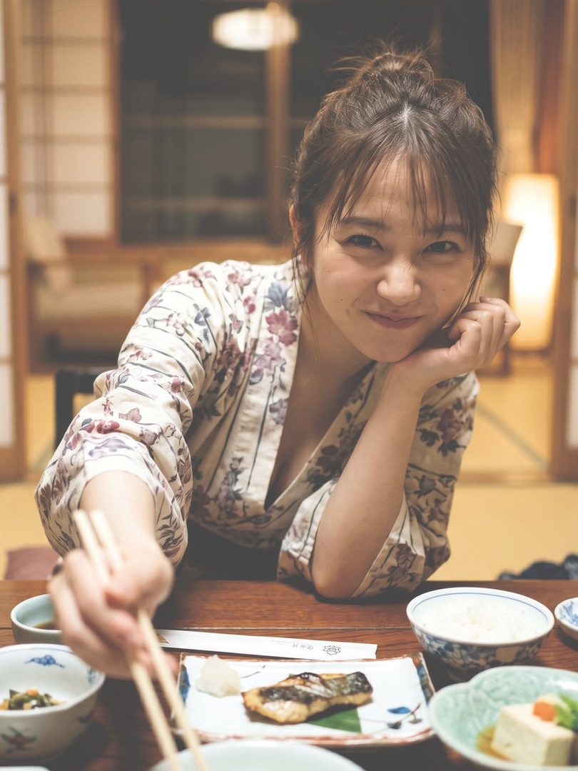 Young Japanese woman in a floral kimono smiling while eating a traditional Japanese meal at a wooden table indoors.