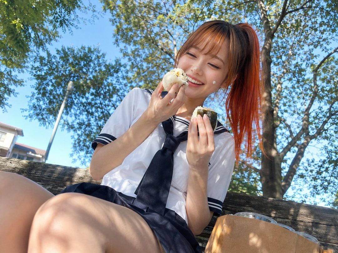 Young woman with orange ponytail hair in sailor uniform happily eating onigiri on a sunny park bench.