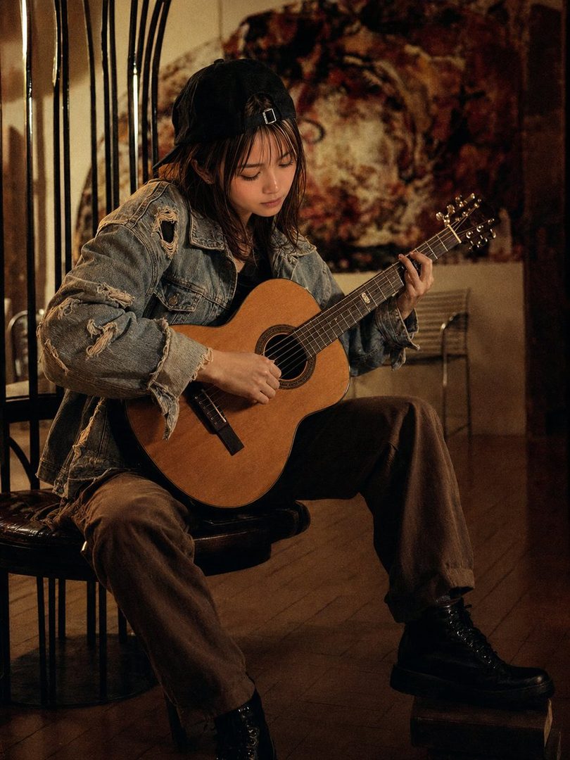 Young woman in a black cap and ripped denim jacket playing an acoustic guitar in a warmly lit studio setting.