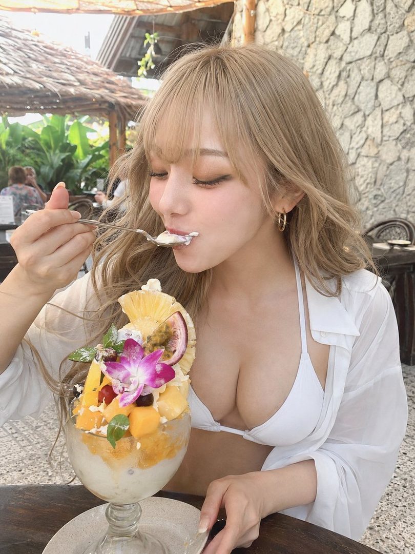 Blonde woman in a white bikini and sheer shirt eating a tropical fruit dessert at an outdoor cafe.