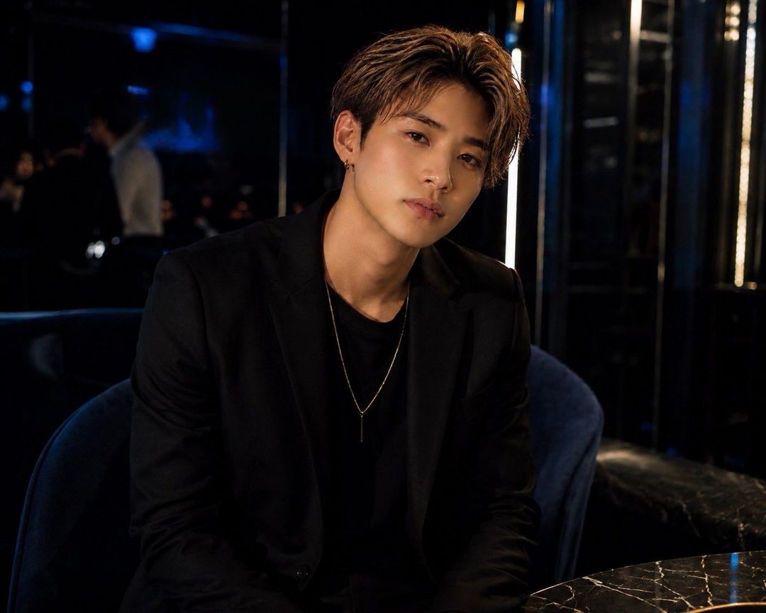 A young man with brown hair and a black blazer sits in a dimly lit nightclub, looking directly at the camera.